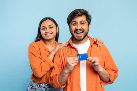 Téléchargez les photos : Couple indien joyeux en chemises orange tenant une carte de crédit, suggérant des achats ou un paiement sur bleu - en image libre de droit