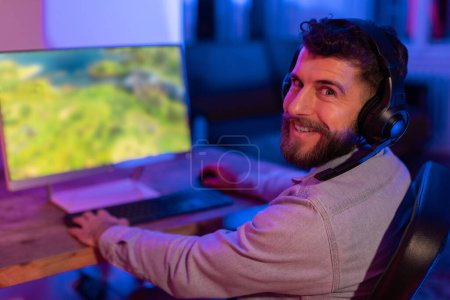 Lächelnder junger bärtiger Mann bewundert lebhafte Spielgrafik auf seinem Computermonitor in einem neonbeleuchteten Raum