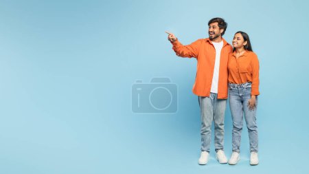 Excited Indian couple pointing to the side while looking in the same direction, against a blue background