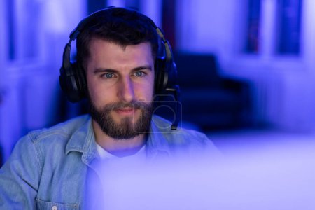 Foto de Hombre tranquilo con un auricular iluminado por la luz azul calma en frente de un monitor de ordenador, el interior del hogar, primer plano - Imagen libre de derechos
