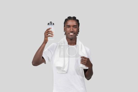 Téléchargez les photos : Un homme afro-américain avec une construction athlétique souriant, tenant une bouteille d'eau de sport, et drapant une serviette sur son épaule, isolé sur un fond uni - en image libre de droit
