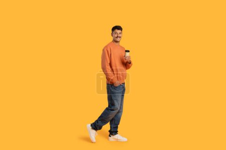 Photo for Funny millennial guy with a moustache carrying a coffee cup while walking, looking relaxed, on an isolated yellow background - Royalty Free Image