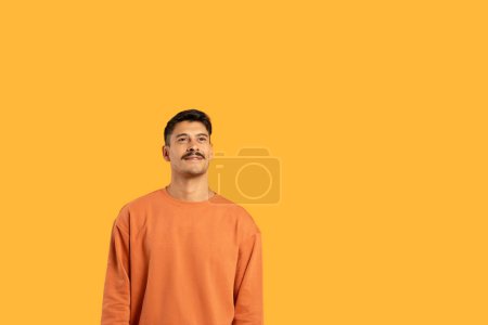 Photo shows a millennial guy with a moustache, standing against a vivid orange isolated background, giving a funny expression, copy space