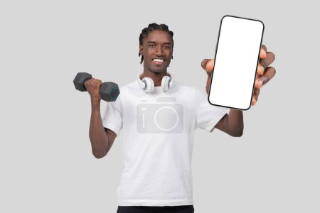 Téléchargez les photos : Un jeune homme afro-américain en tenue de sport tient un haltère dans une main et présente un écran de smartphone vierge avec l'autre, sur un fond gris - en image libre de droit