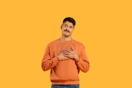 Photo for A young man with a mustache, wearing an orange sweater, smiles and makes a heart gesture with his hands against a solid yellow background - Royalty Free Image