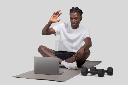 Ein schwarzer, athletischer Mann ist mit einem Videoanruf auf seinem Laptop beschäftigt, während er auf einer Gymnastikmatte sitzt, mit Hanteln in der Nähe, isoliert auf weiß eingefangen