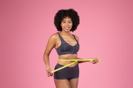 A confident sporty African American lady holds a measuring tape around her waist on an isolated pink background, representing health and fitness