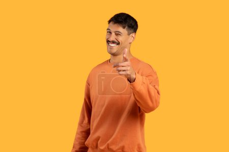 Freundlicher Millennial-Typ mit Schnurrbart, der mit einem lustigen Lächeln in die Kamera zeigt, vor einem lebhaften orangefarbenen Hintergrund stationiert