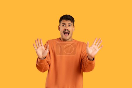 Photo for A young man in an orange sweatshirt stands with raised hands expressing surprise or shock against a vivid orange background - Royalty Free Image