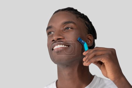 A close-up of a cheerful young African American man with braided hair, shaving his face with a blue disposable razor against a white background