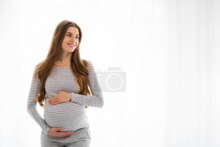 A content pregnant woman in a striped shirt and grey pants holds her belly and looks to the side with a gentle smile, copy space