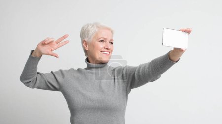 Ältere europäische Seniorin posiert mit Smartphone und blinkt ein Friedenszeichen, das die s3niorlifes Umarmung jugendlicher Trends verkörpert