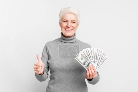 Téléchargez les photos : Femme européenne âgée souriante donnant un coup de pouce avec de l'argent en main, suggérant la confiance financière dans s3niorlife - en image libre de droit