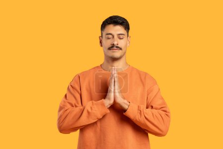 Millennial guy with a moustache in a prayer pose against a funny isolated orange background, radiating tranquility and focus