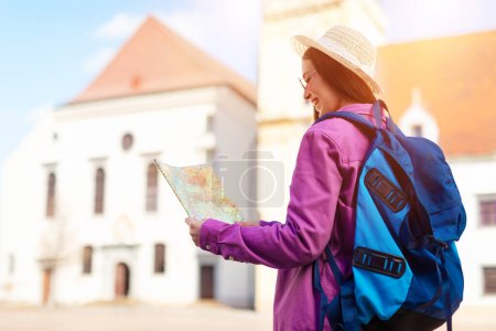 Vista trasera de la mujer viajera con un mapa frente a una iglesia iluminada por el sol, que transmite una sensación de aventura