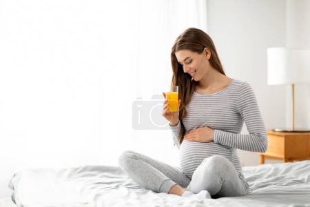 Téléchargez les photos : Une femme enceinte rayonnante s'assoit confortablement sur un lit, berçant sa bosse de bébé et souriant tout en tenant un verre de jus d'orange - en image libre de droit