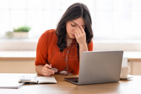 Eine fokussierte Frau aus dem Mittleren Osten in einer leuchtend orangefarbenen Bluse arbeitet fleißig an ihrem Laptop an einem Holztisch und notiert Notizen in ein Notizbuch.