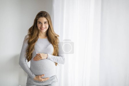 Photo for A happy and radiant pregnant woman with long hair in cozy clothing is standing by a white curtain, cradling her belly - Royalty Free Image