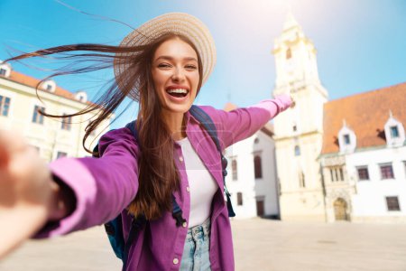 A joyful woman tourist in a straw hat takes a playful selfie with historic European buildings in the background