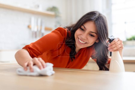 Joyful middle eastern woman using a cleaning spray to wipe down a wooden kitchen counter in bright daylight