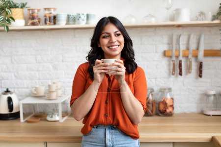 Téléchargez les photos : Belle femme du Moyen-Orient avec un sourire chaud tenant une tasse de café dans une cuisine confortable à la maison - en image libre de droit