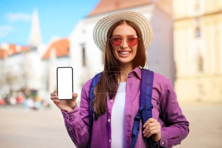 Fröhliche junge Reisende zeigt leere Smartphone-Bildschirm-Attrappe mit historischem Stadtbild hinter sich