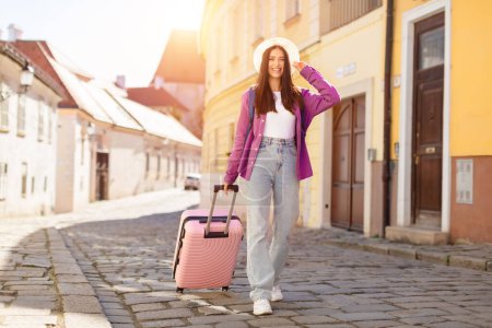 Téléchargez les photos : Une jeune voyageuse vêtue d'une veste violette et d'un jean bleu sourit en marchant dans une rue pavée avec sa valise rose dans une ville pittoresque - en image libre de droit