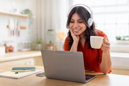 Téléchargez les photos : Jeune femme du Moyen-Orient avec écouteurs utilisant un ordinateur portable et profiter d'une tasse de café, incarne multitâche à la maison - en image libre de droit