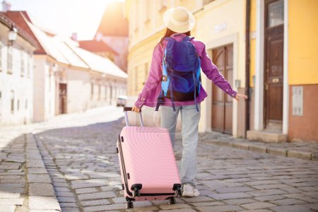 Téléchargez les photos : Femme voyageuse avec valise rose errant dans une rue pavée ensoleillée dans une ville européenne pittoresque, incarnant l'aventure et la découverte, vue de dos - en image libre de droit