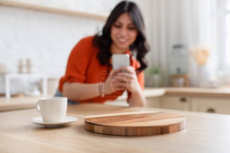 Foto de El foco está en una taza de café blanco con una mujer borrosa usando un teléfono inteligente en el fondo en una cocina - Imagen libre de derechos