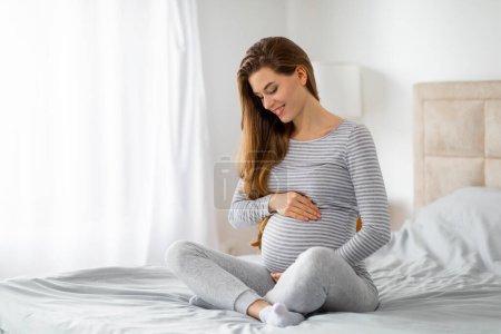Téléchargez les photos : Une femme enceinte souriante en vêtements rayés s'assoit confortablement sur son lit, caressant son ventre avec une expression sereine - en image libre de droit