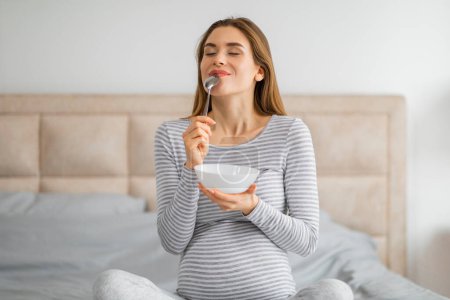 Foto de Una joven embarazada vestida a rayas está disfrutando del desayuno en la cama, lamiendo una cuchara con una expresión satisfecha - Imagen libre de derechos