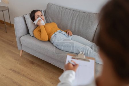 Eine junge Frau liegt auf einer Couch und wirkt gestresst, während sie mit ihrem Therapeuten sitzt, Konzept für psychische Gesundheit
