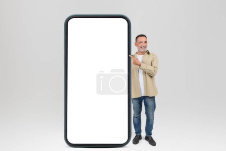 Photo for Cheerful elderly man gestures towards a blank large smartphone screen, isolated on white background, mockup, copy space - Royalty Free Image