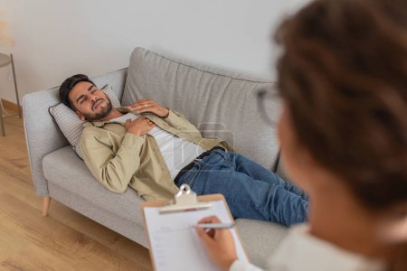 Eine Therapiesitzung ist im Gange, bei der ein junger Mann auf dem Sofa über seine Ehe diskutiert, während ein Therapeut Notizen macht.