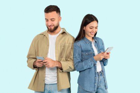 Foto de Un hombre y una mujer adultos jóvenes se paran uno al lado del otro, absortos en sus teléfonos inteligentes sobre un fondo azul, lo que sugiere una conectividad moderna - Imagen libre de derechos