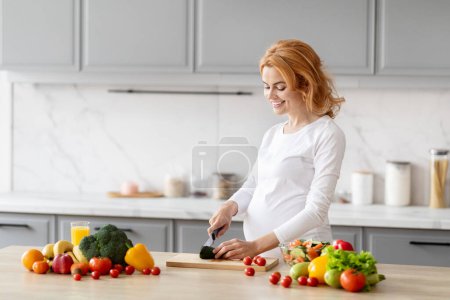 Foto de Una mujer embarazada alegre está parada en una cocina moderna, cortando verduras en una tabla de cortar, rodeada de productos frescos - Imagen libre de derechos