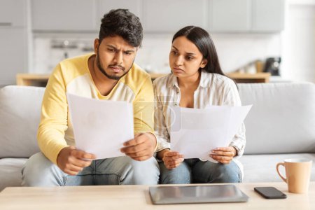 Photo for This eastern millennial couple looks closely at financial documents, displaying expressions of concern, possibly reviewing bills or statements at home - Royalty Free Image