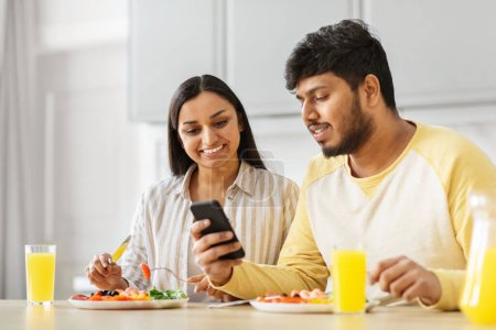 Téléchargez les photos : Un jeune couple indien dans la cuisine examine un smartphone, dépeignant des milléniaux fusionnant la technologie avec leur amour et leur vie quotidienne - en image libre de droit