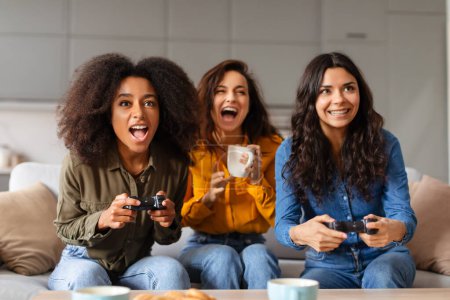 Téléchargez les photos : Trois jeunes filles multiraciales appréciant les jeux vidéo ensemble, exprimant l'excitation dans un environnement familial - en image libre de droit