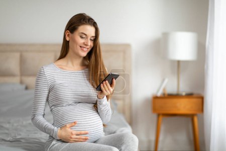 Téléchargez les photos : Une future mère heureuse assise sur le lit en regardant son téléphone portable, touchant doucement son ventre, dans une pièce bien éclairée - en image libre de droit