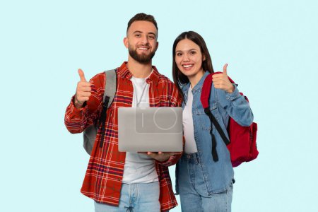 Feliz pareja de estudiantes con mochilas y un ordenador portátil que da pulgares hacia arriba, en un fondo de estudio azul fresco