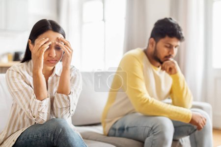 An upset Indian woman sits with her head in her hands beside a man who looks frustrated, both seated on a couch, indicating relationship issues
