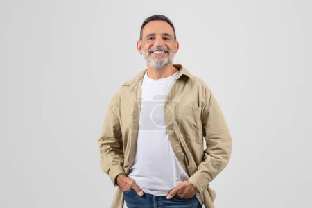 Retrato de un anciano alegre, luciendo chaqueta y jeans, las manos descansadas en sus bolsillos, aisladas sobre un fondo blanco