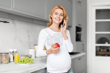 Téléchargez les photos : Une heureuse femme enceinte européenne parle au téléphone dans sa cuisine, entourée d'éléments nutritionnels de cuisine - en image libre de droit