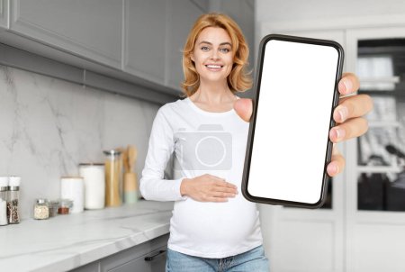 Mujer embarazada europea en una cocina que presenta una pantalla de teléfono en blanco, mostrando la mezcla de tecnología moderna y salud prenatal