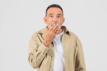 Mann mittleren Alters mit beigem Hemd wirkt geschockt, die Hand über dem Mund vor weißem Hintergrund