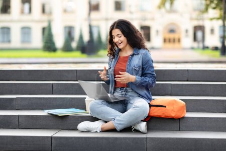 Une femme de l'Est concentrée engagée avec son ordinateur portable, assise dans les escaliers d'un campus un cadre d'éducation moderne