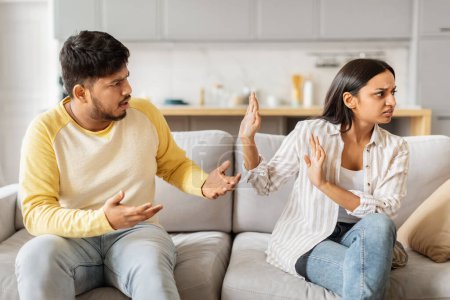 An adult Indian man and woman are seen having an emotional argument while sitting on a sofa in a well-lit living room setting