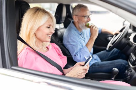 Téléchargez les photos : Une femme mûre vérifie son téléphone dans le siège passager pendant que l'homme mange et conduit, à la fois contenu et à l'aise dans un véhicule moderne - en image libre de droit